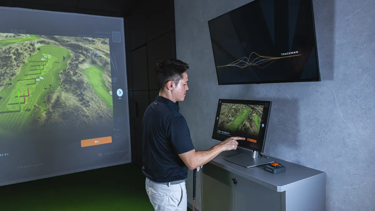 T-One Golf Studioの打席でタッチパネルを操作してコースを選ぶゴルファー スクリーンにコースの全景が映っている